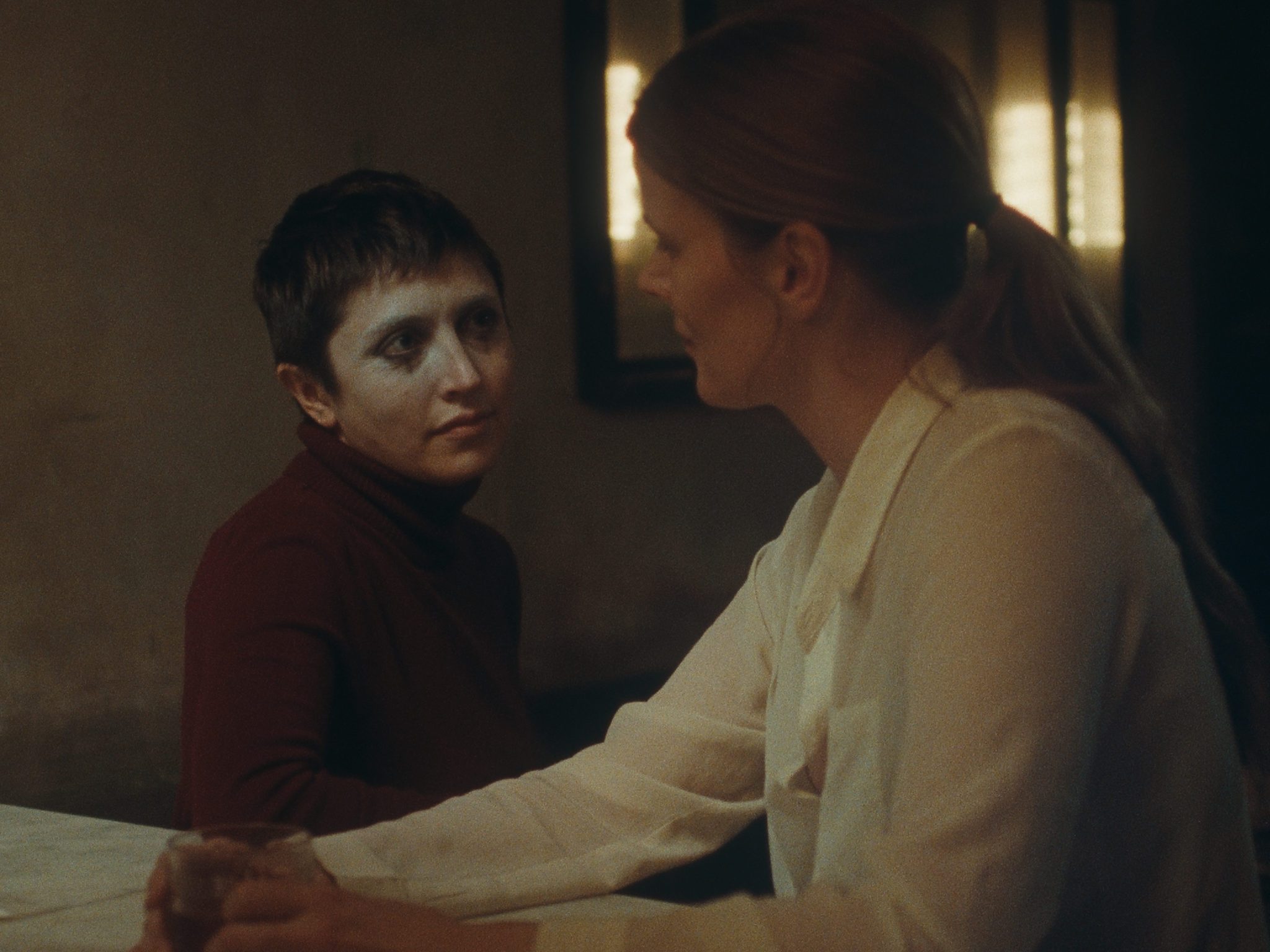 The protagonist's face is only partially covered in makeup as they sit at table, they appear to be engaged in a tense discussion with a woman.